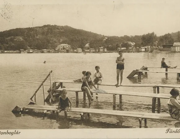 Das Bild zeigt eine historische Postkarte des Plattensees mit Personen, die in Badekleidung auf Stegen sitzen oder stehen.