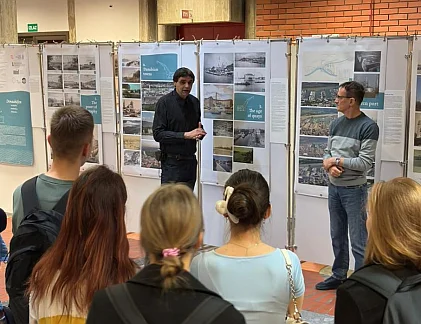 Das Bild zeigt 2 Männer vor Ausstellungstafeln, die zu einer Menge junger Menschen sprechen.