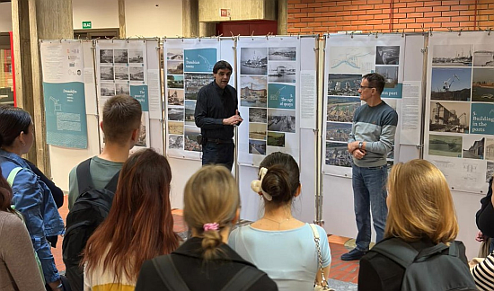 Das Bild zeigt 2 Männer vor Ausstellungstafeln, die zu einer Menge junger Menschen sprechen.